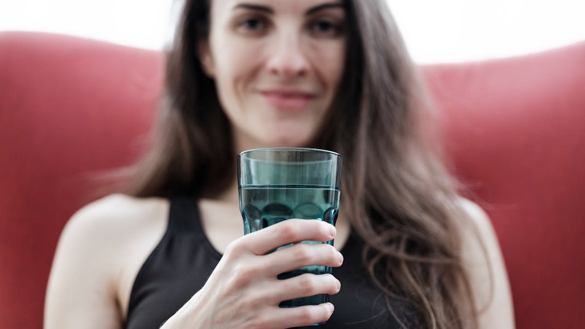 Smiling woman with long hair holding a glass of water, seated indoors with a red chair.